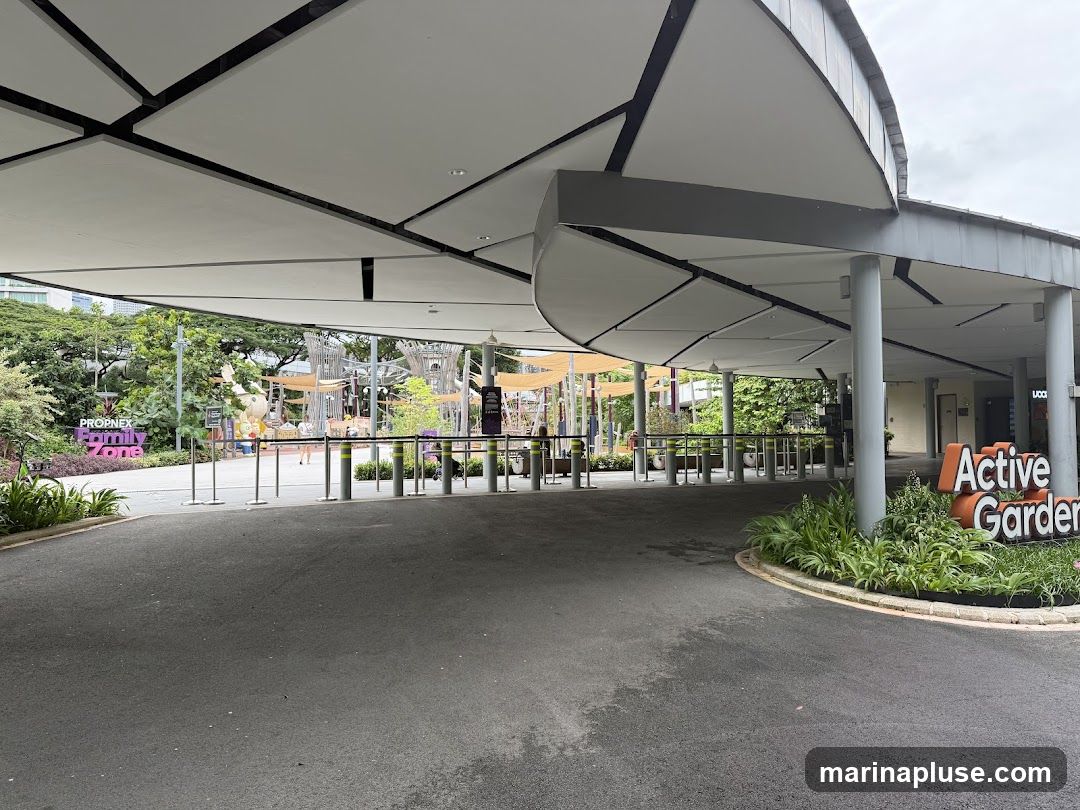 A wide shot of the entrance to an outdoor attraction. A large, modern canopy covers a paved walkway leading to turnstiles. To the left, a sign reads "PROPNEX Family Zone." To the right, a prominent sign says "Active Garden." In the background, there are various structures and greenery, suggesting a park or recreational area.