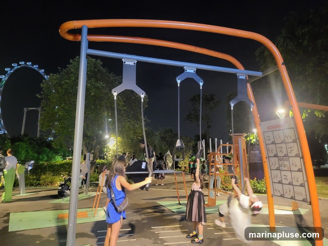 A playground scene at night with children playing on swings and other equipment. In the background, a large Ferris wheel is illuminated. There are trees and park lighting visible. A sign with exercise instructions is mounted on the orange structure.