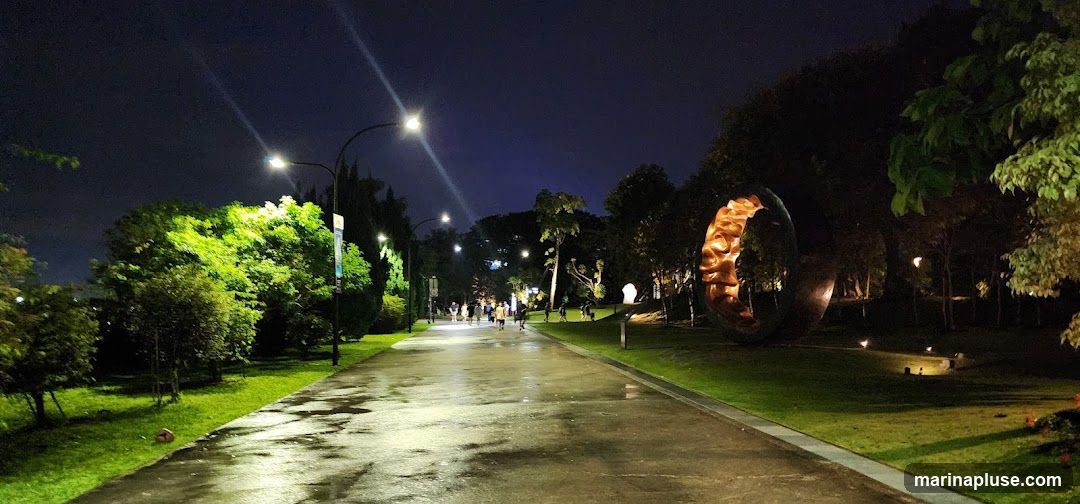 A wet pathway in a park at night, illuminated by streetlights. Lush green trees line the path, and a large, abstract sculpture with a glowing interior is visible on the right. Several people are walking along the path in the distance.
