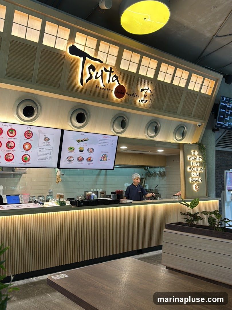 A view of the interior of a Tsuta Japanese Soba Noodles restaurant. The restaurant's logo is prominently displayed above the counter. Digital menus showing ramen, rice, sides, and drinks are visible. A staff member is seen behind the counter. A sign on the right reads "The World's First Michelin Star Ramen".