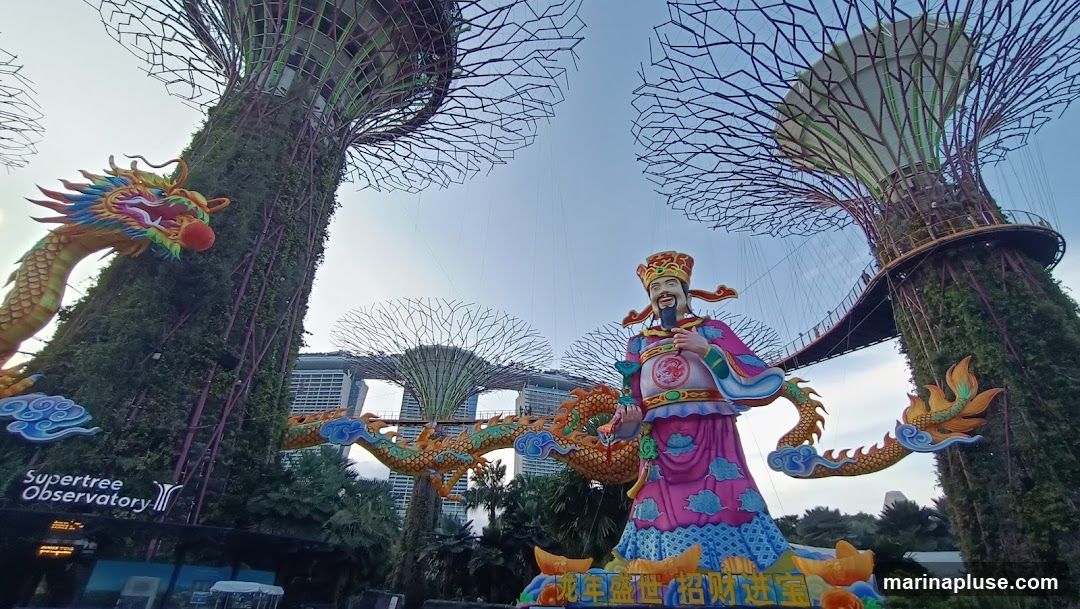 A vibrant display of Chinese New Year decorations featuring a large statue of a traditional Chinese deity, flanked by colorful dragon figures. The Supertree Observatory is visible in the background, with its distinctive tree-like structures and skywalk. The decorations appear to be part of a festive event, possibly celebrating the Lunar New Year.