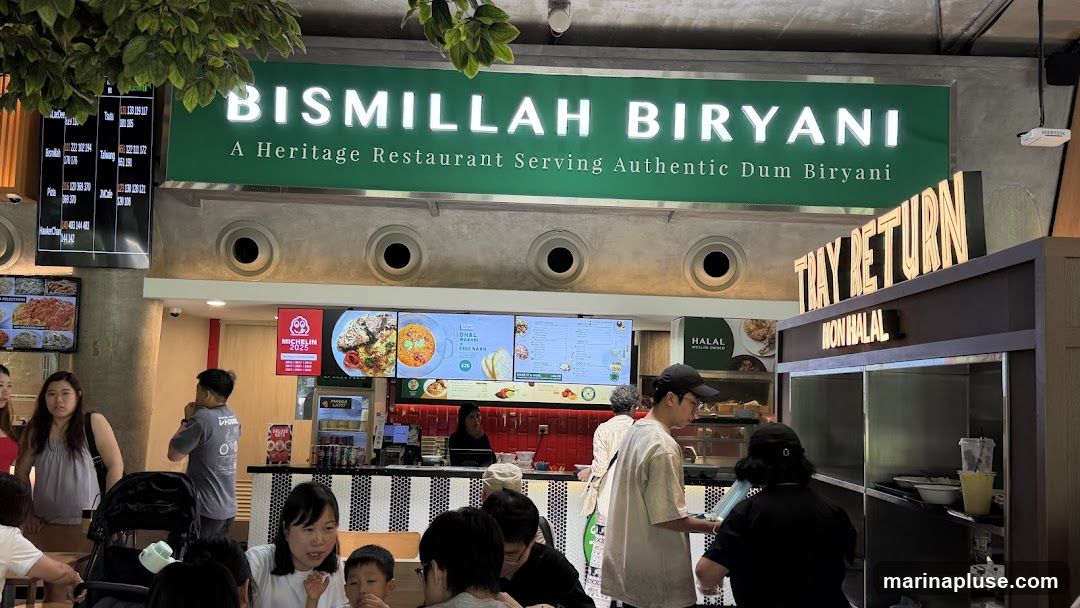 A busy restaurant interior with a large sign that reads "BISMILLAH BIRYANI A Heritage Restaurant Serving Authentic Dum Biryani". Below the sign are digital menu boards displaying food items and prices. People are seen ordering and dining. A "TRAY RETURN" sign is visible above a counter labeled "NON HALAL".