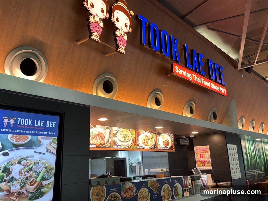 A view of the interior of a Thai food restaurant called Took Lae Dee. The restaurant's name is displayed prominently on a wooden wall with blue lettering and a red sign that says "Serving Thai Food Since 1972". There are images of Thai dishes displayed, including Green Curry and Pad Thai. A digital screen on the left shows more food images and the restaurant's name and slogan.