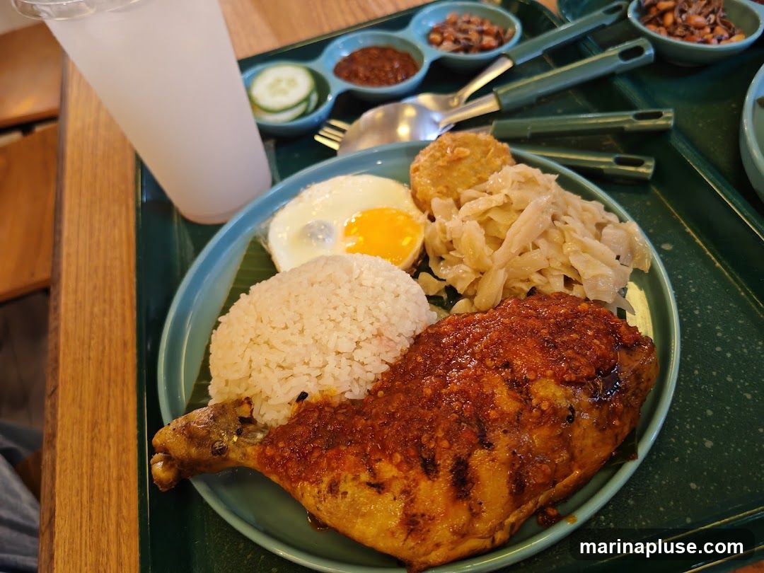 A close-up shot of a meal served on a teal tray. The meal includes a generous portion of white rice, a piece of grilled chicken leg covered in a spicy red sauce, a fried egg, and a side of shredded vegetables. There are also small dishes containing condiments, including sliced cucumbers and chili paste. A tall, frosted glass of a beverage is visible in the upper left corner.