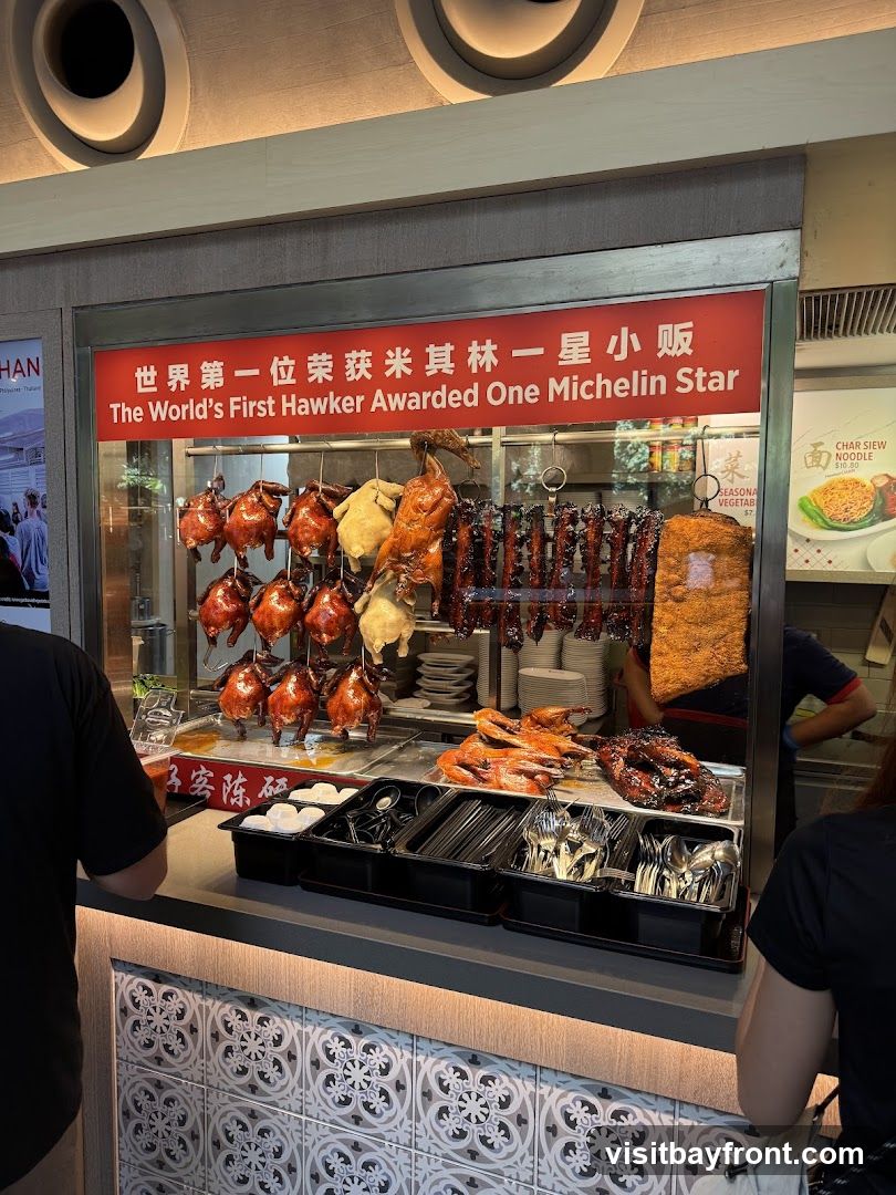A stall displaying various roast meats, including whole chickens and ducks, hanging from hooks. Below, trays hold prepared meats and cutlery. A sign above proclaims "The World's First Hawker Awarded One Michelin Star." Another sign advertises "Char Siew Noodle" for $10.00.