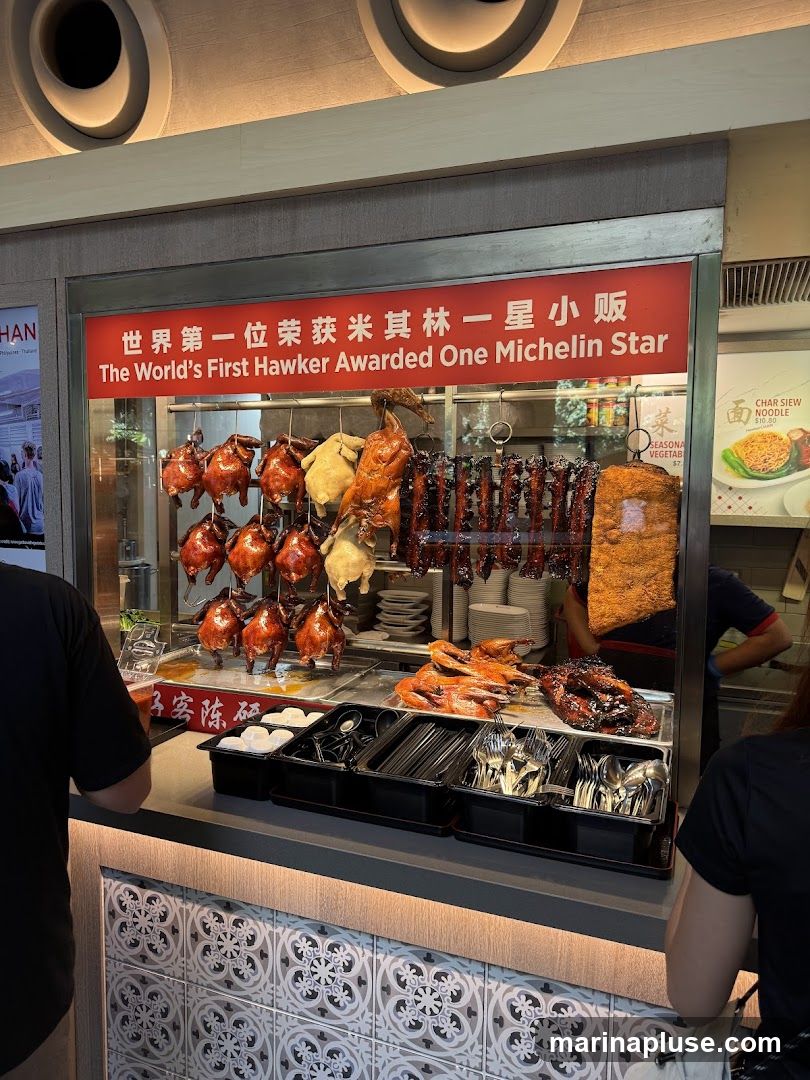 A stall displaying various roast meats, including whole chickens and ducks, hanging from hooks. Below, trays hold prepared meats and cutlery. A sign above proclaims "The World's First Hawker Awarded One Michelin Star." Another sign advertises "Char Siew Noodle" for $10.00.