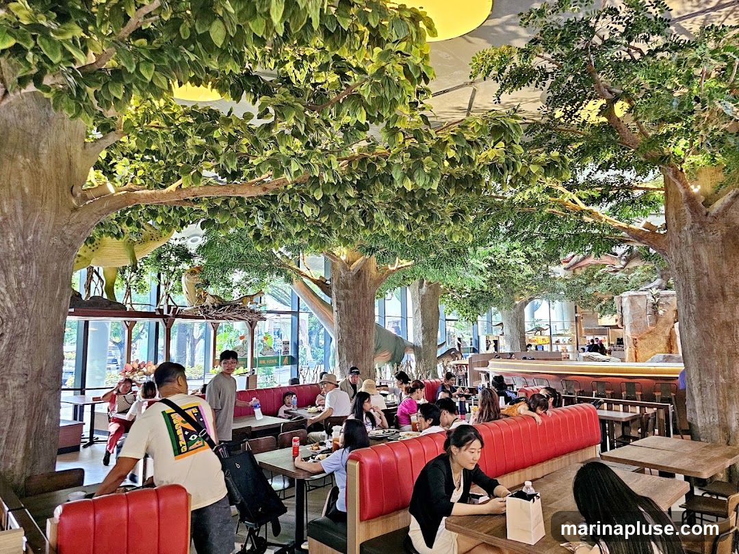 The interior of a restaurant designed with a prehistoric theme, featuring large artificial trees with lush green foliage, and dinosaur models. Several patrons are seated at tables, some dining and others interacting. The seating includes red booths and wooden tables. The overall ambiance is lively and family-friendly, with a focus on creating an immersive experience.