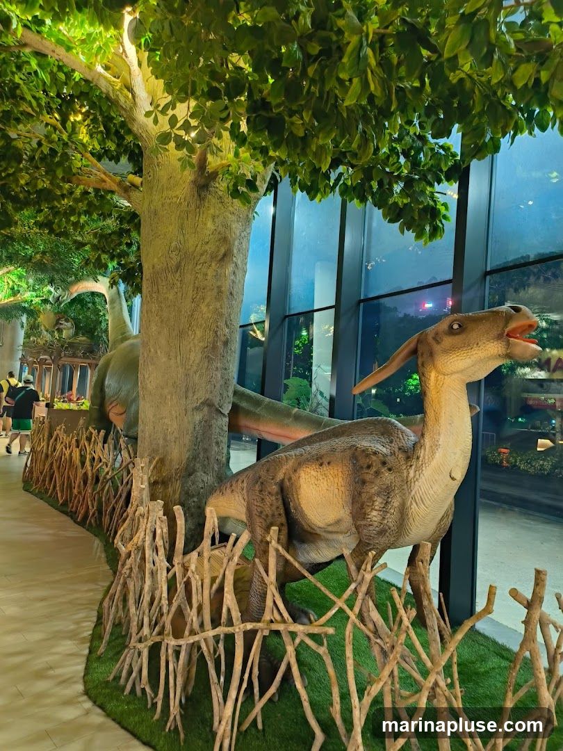 A display of realistic dinosaur models, including a Parasaurolophus in the foreground and a Brachiosaurus in the background, set against a backdrop of large windows overlooking a city at night. The exhibit features artificial trees, greenery, and a rustic wooden fence.