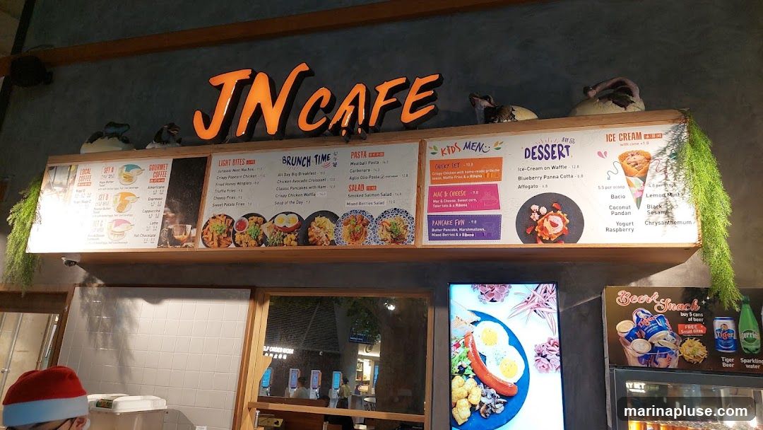 A menu board for JN Cafe displays various food and drink options, including light bites, brunch items, pasta, salads, kids' meals, desserts, and ice cream flavors. The menu is mounted on a wall above a counter, with decorative elements like dinosaur figurines and hanging plants.
