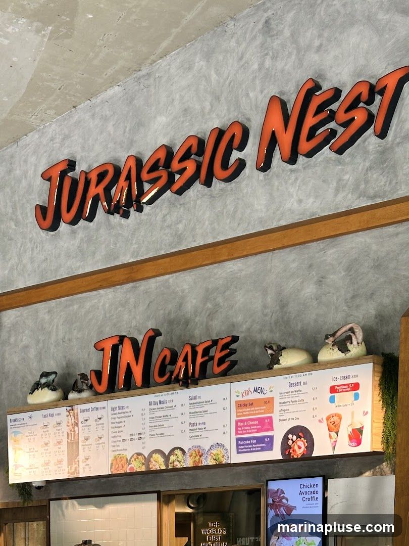 A wall-mounted menu board for Jurassic Nest Cafe, featuring sections for Breakfast, Local Kopi, Gourmet Coffee, Light Bites, All Day Meals, Salad, Pasta, Kids Menu, and Desserts. The menu has images of food items and prices.