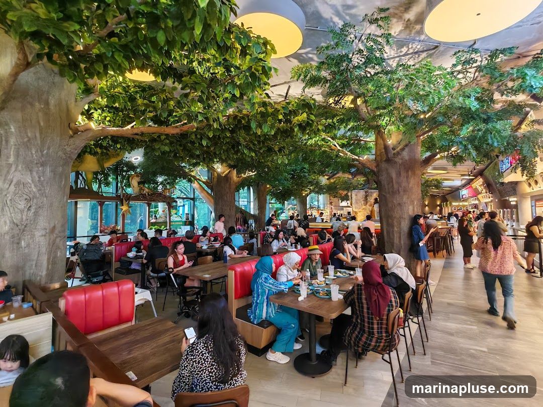 A bustling food court with a unique theme, featuring large artificial trees with lush green foliage, creating a canopy over the dining area. Several patrons are seated at wooden tables with red booth seating, enjoying their meals. In the background, various food stalls are visible, with some signage indicating different eateries. The overall ambiance is lively and family-friendly, with a focus on creating an immersive dining experience.