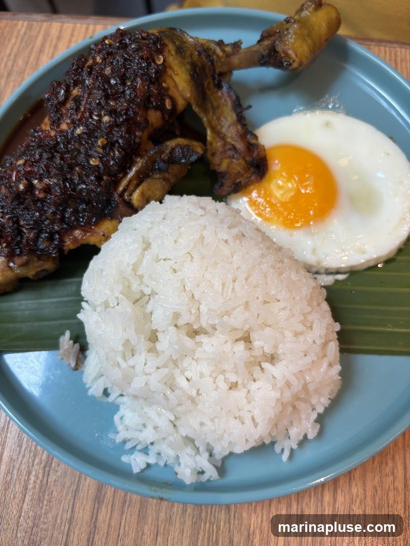 A close-up shot of a plate of food. The plate contains a portion of white rice, a fried egg with a runny yolk, and a piece of chicken leg quarter. The chicken is coated in a dark, textured sauce with visible chili flakes. A green banana leaf is visible beneath the rice and egg. The plate is a muted teal color and sits on a wooden surface.