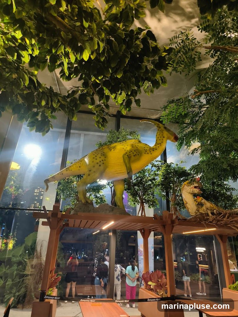 An indoor space decorated with artificial trees and dinosaur models. A large yellow dinosaur stands on a platform, while a smaller dinosaur is nestled in a nest. People are visible through a glass partition, and tables and chairs are set up in the foreground. The signage "Jurassic Nest" is visible on several elements within the space.