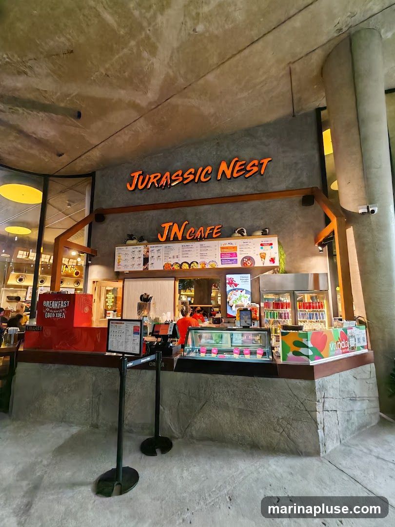 The exterior of a cafe with a concrete facade. The cafe's name, "JURASSIC NEST JN CAFE", is displayed in large orange lettering above the counter. Below the name, a menu board is visible, along with display cases filled with food items. The counter area is well-lit, and there are refrigerators stocked with drinks.