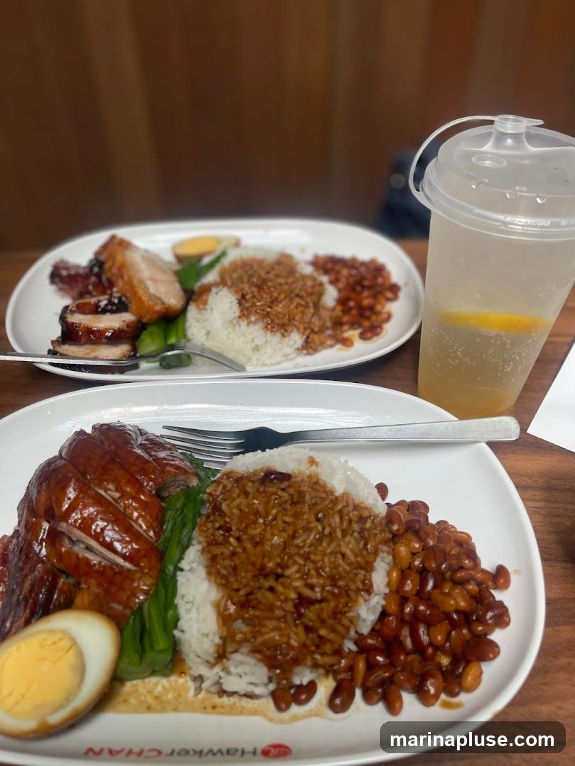 Two plates of rice with roasted meats and a drink. The plate in the foreground features sliced roast duck, a hard-boiled egg, green vegetables, rice topped with gravy, and peanuts. The plate in the background appears to have char siu (barbecued pork) and crispy pork belly, also served with rice, peanuts, and a hard-boiled egg. A clear plastic cup with a straw, containing a light-colored beverage with a lemon slice, is visible on the right. The text "HAWKER CHAN" is visible on the rim of the plate in the foreground.