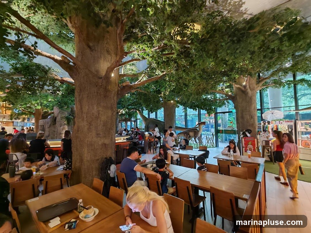 The interior of a restaurant designed with a jungle theme, featuring large artificial trees with lush foliage. Several dinosaur models are visible throughout the space, including a large one in the background. The restaurant is filled with patrons dining at wooden tables. The overall ambiance is lively and family-friendly, with a retail display area visible on the right side of the image.