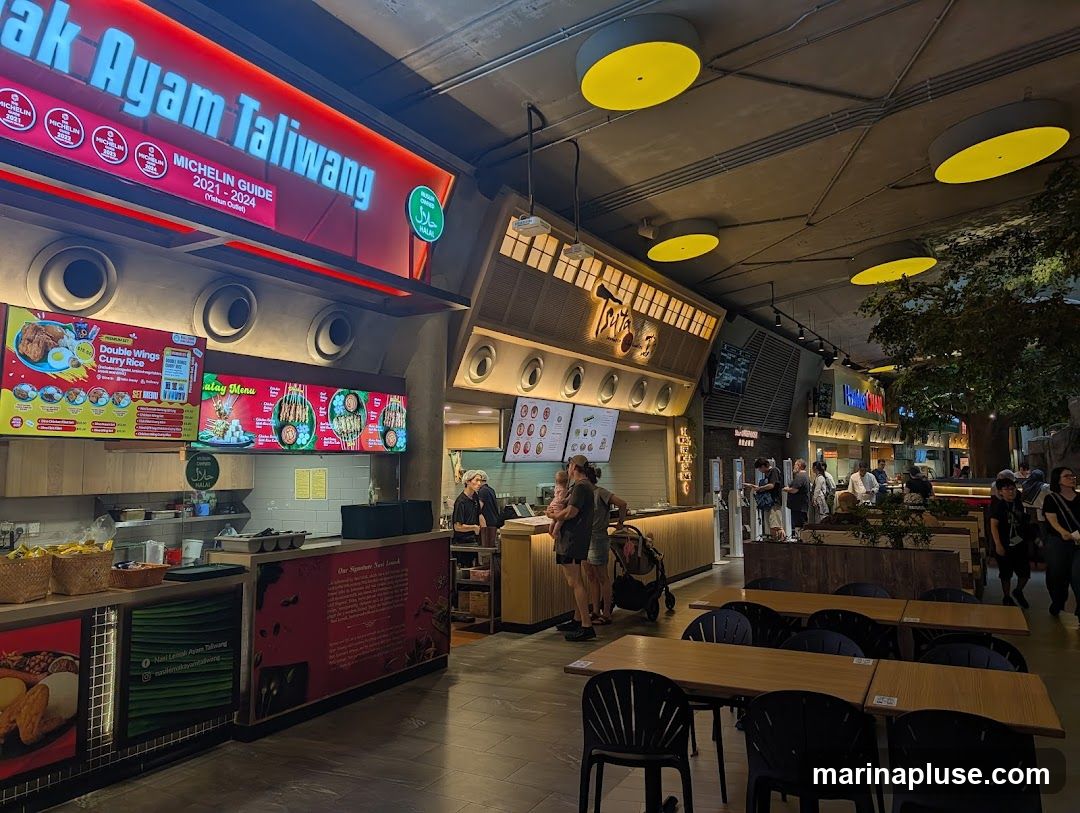 A bustling food court with various stalls and seating areas. One stall prominently displays "Taliwang" and "Michelin Guide" accolades. Menus with food items and prices are visible, along with people ordering and dining.