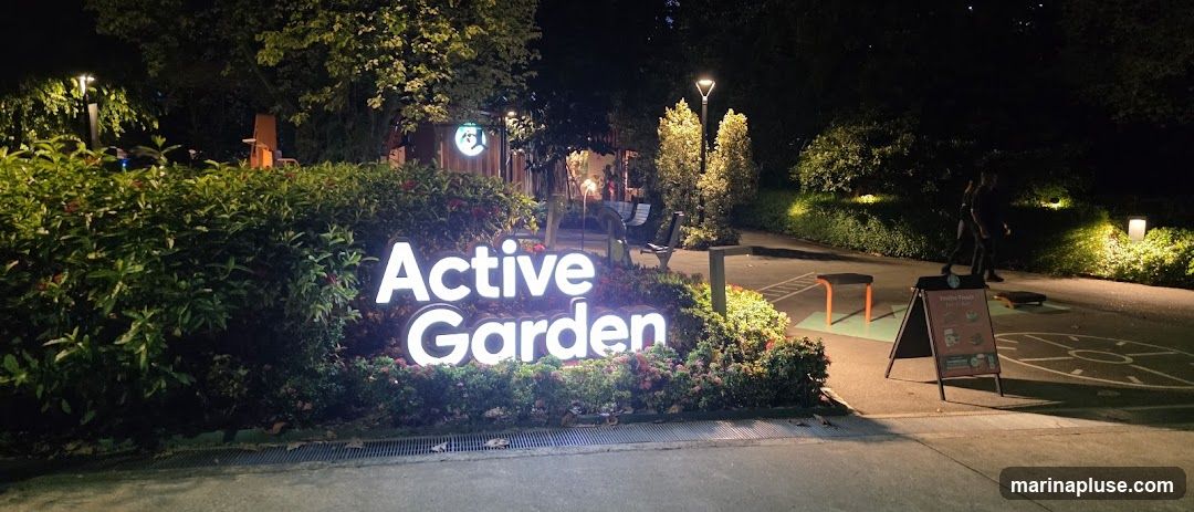 A large, illuminated sign reads "Active Garden" in white lettering, set against a backdrop of lush green bushes and trees at night. In the background, there are outdoor exercise equipment, benches, and lighting. A-frame sign is visible on the right.