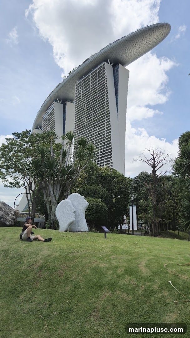 A person sits on a grassy hill in front of the Marina Bay Sands hotel in Singapore. A large, abstract sculpture of two faces is visible in the foreground. The sky is partly cloudy.