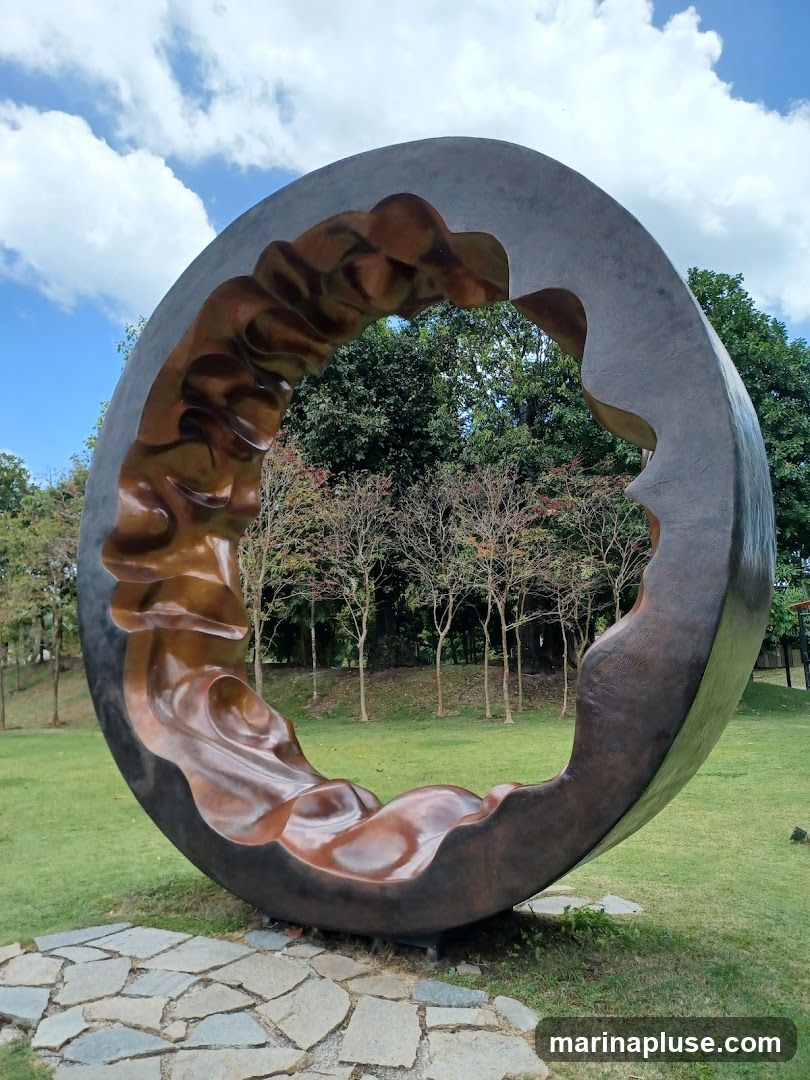A large, abstract sculpture with a dark, textured outer ring and a polished, bronze-colored interior with flowing, wave-like patterns. The sculpture is situated outdoors on a grassy lawn, with a stone pathway leading up to its base. In the background, there are trees and a bright blue sky with scattered clouds.