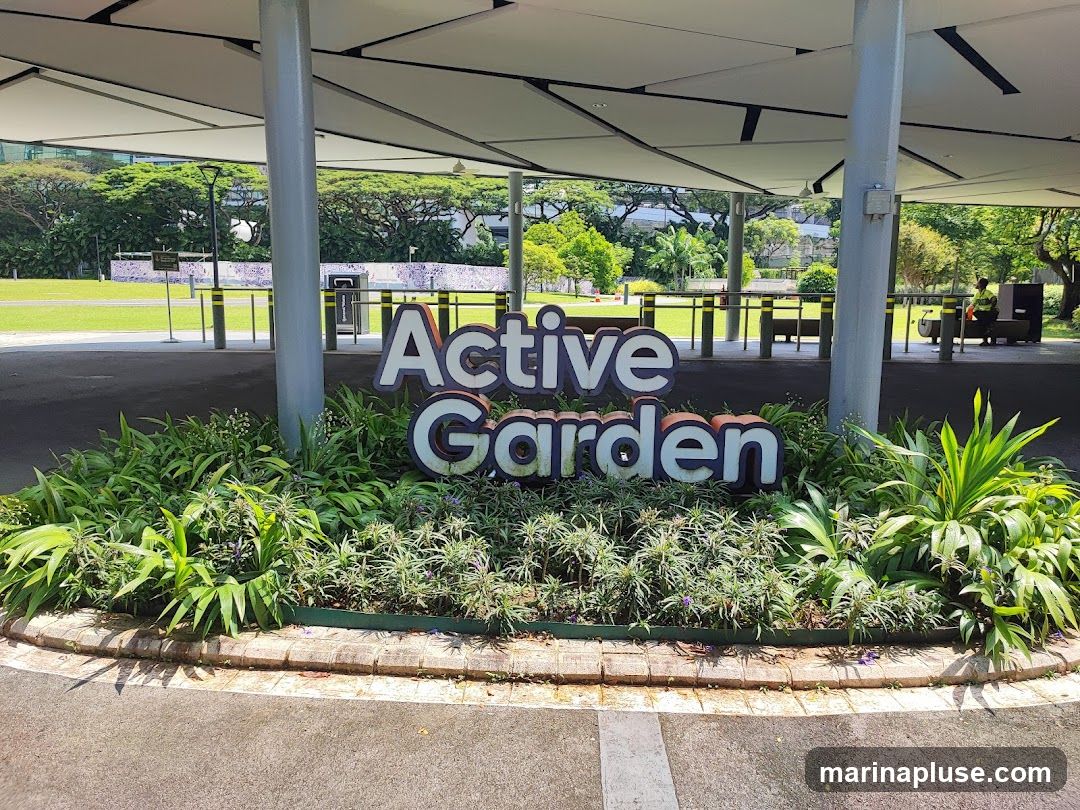 A large, three-dimensional sign spelling out "Active Garden" is prominently displayed in front of lush greenery. The sign has white letters with blue outlines for "Active" and white letters with orange outlines for "Garden." Behind the sign, a covered walkway with a modern, angular roof supported by pillars extends outwards. Beyond the walkway, a grassy field is visible, with trees and buildings in the distance. A person in a yellow vest is seen in the background near some equipment.