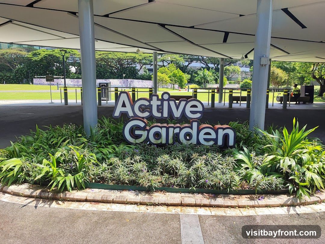 A large, three-dimensional sign spelling out "Active Garden" is prominently displayed in front of lush greenery. The sign has white letters with blue outlines for "Active" and white letters with orange outlines for "Garden." Behind the sign, a covered walkway with a modern, angular roof supported by pillars extends outwards. Beyond the walkway, a grassy field is visible, with trees and buildings in the distance. A person in a yellow vest is seen in the background near some equipment.