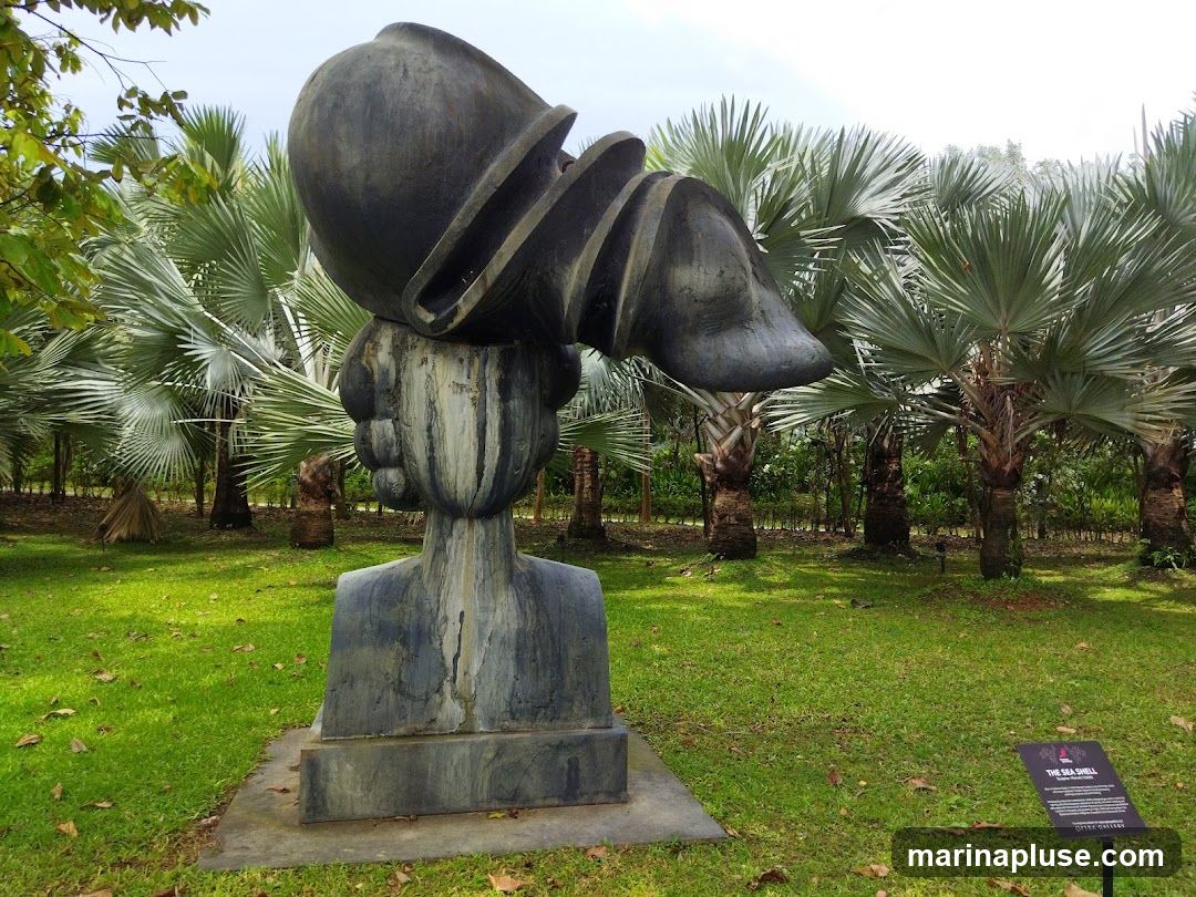 A large, dark gray abstract sculpture resembling a head with a shell-like object on top is displayed in a lush garden. The sculpture is positioned on a concrete base in front of a row of tall palm trees with fan-shaped leaves. The ground is covered in green grass with scattered fallen leaves. A small black sign with white text is placed to the right of the sculpture.