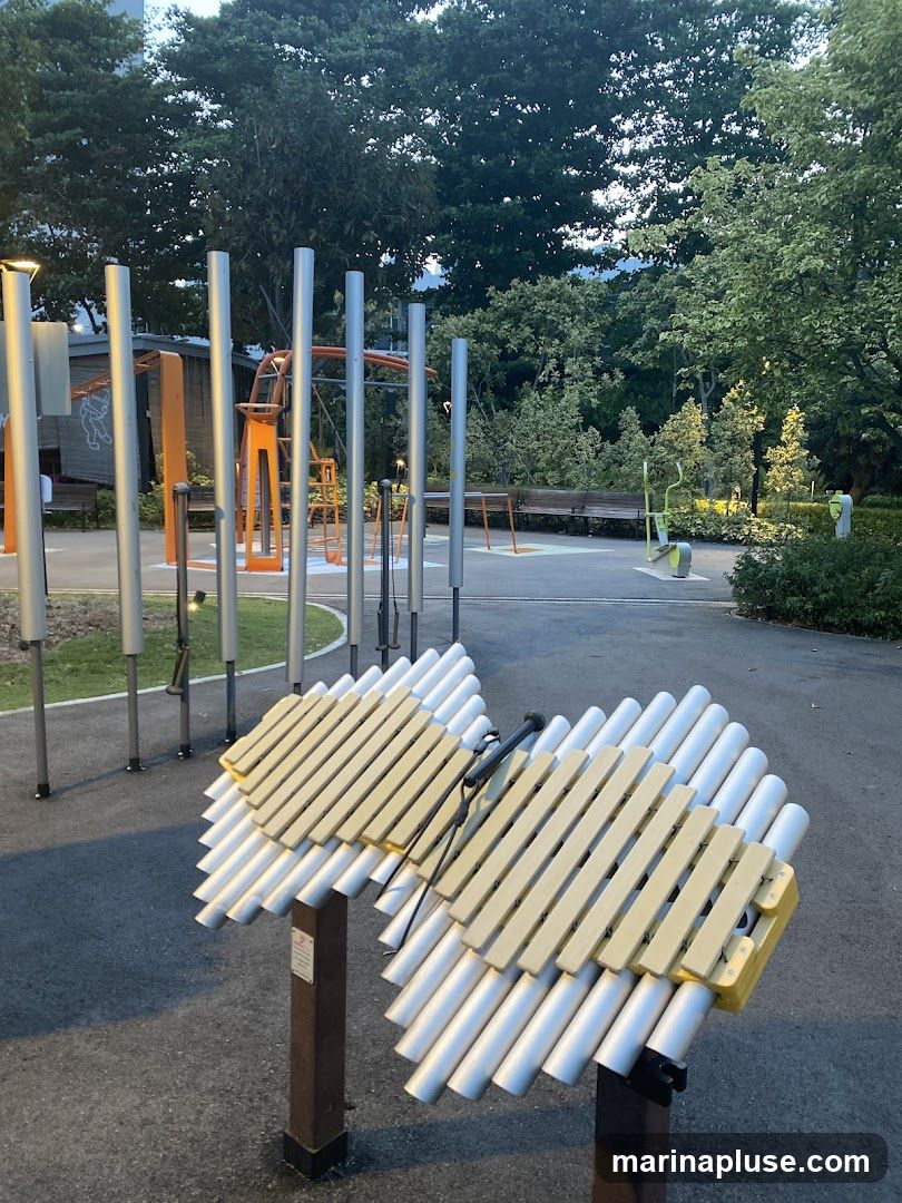 A park setting with various outdoor musical instruments, including a large xylophone-like instrument in the foreground. In the background, there are metal poles and structures that appear to be part of a playground or exercise area. The scene is set amongst trees and greenery, with paved pathways.