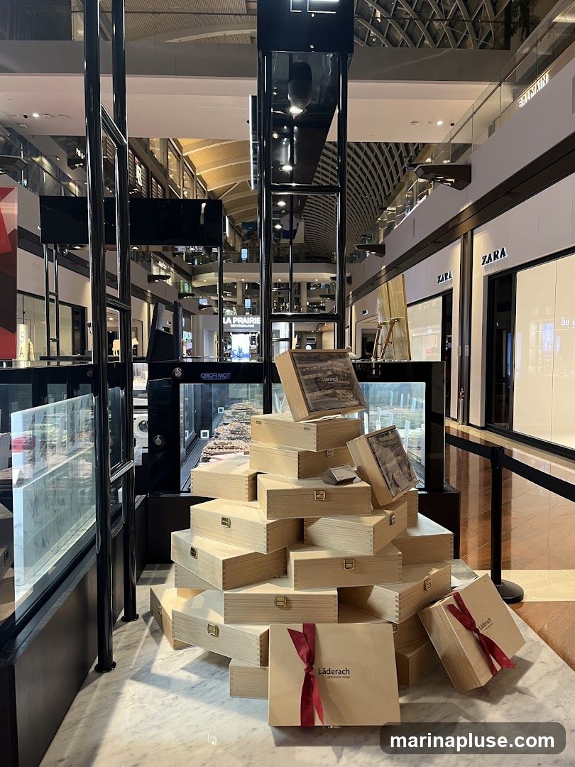 A display of stacked wooden gift boxes with red ribbons, some featuring the brand name "Läderach". The display is set up in a shopping mall, with storefronts like Zara and Balmain visible in the background. A glass display case with products is also visible to the left.