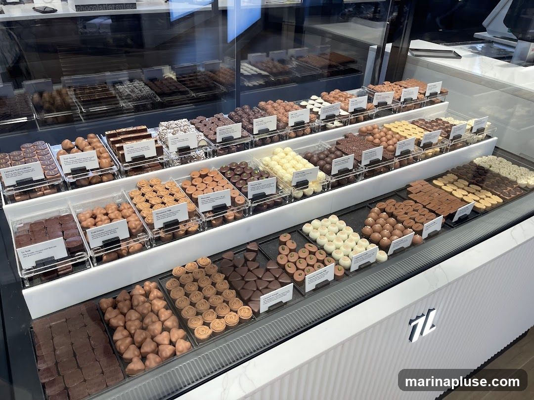 A display case filled with various types of chocolates, including dark, milk, and white chocolate. The chocolates are arranged in clear plastic containers with labels. Some chocolates are square, while others are molded into shapes like hearts and rosettes. The display is well-lit, showcasing the variety and quality of the confectionery. A logo is visible on the front of the display case.