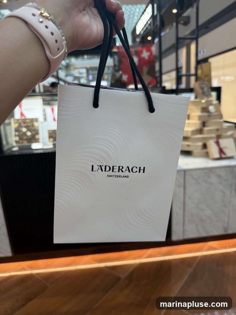 A person holds a white shopping bag with the Läderach Switzerland logo. The background appears to be a shopping mall with displays of chocolates and other merchandise.
