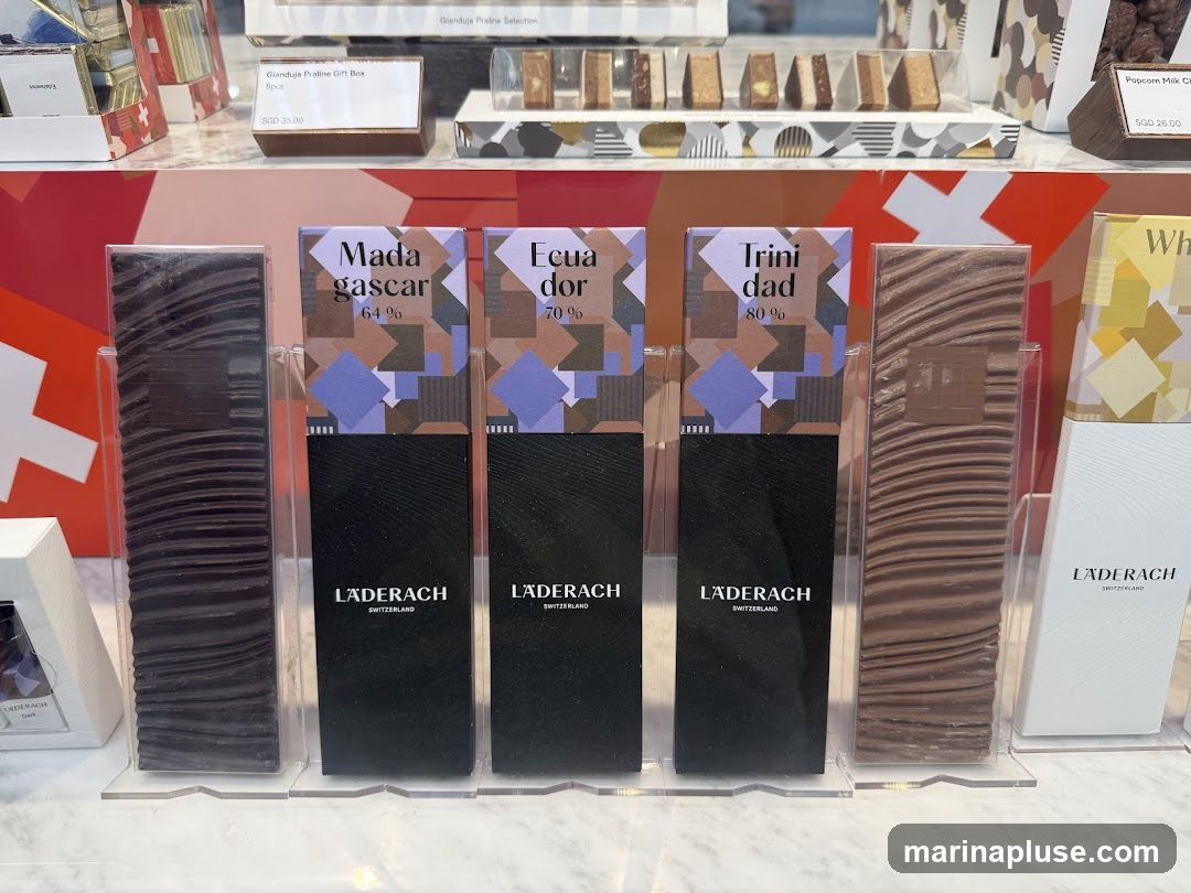 A display of various Läderach chocolate bars and gift boxes. The bars are presented vertically in clear plastic holders, with labels indicating origin and percentage of cocoa. Visible labels include 'Madagascar 64%', 'Ecuador 70%', and 'Trinidad 80%'. In the background, gift boxes are visible, including a 'Gianduja Praline Gift Box' priced at SGD 35.00 and 'Popcorn Milk Choc' priced at SGD 26.00.