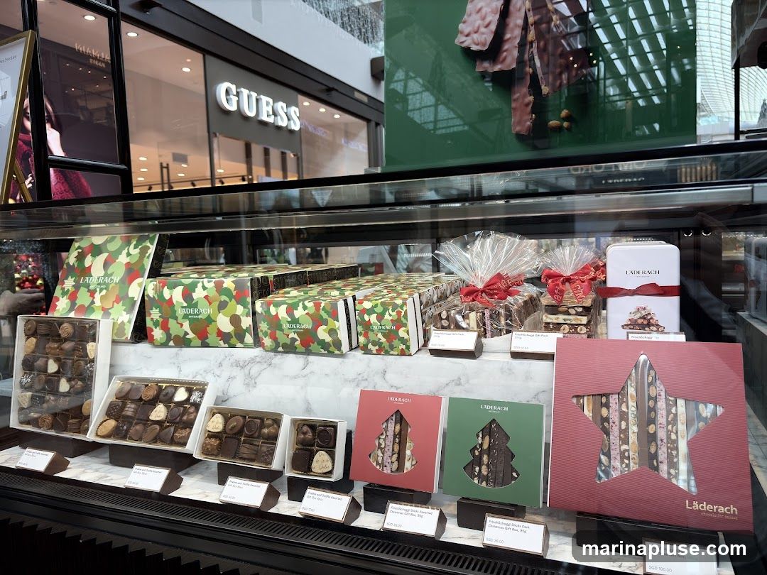 A display case filled with various Läderach chocolates, including gift boxes, assorted chocolates, and chocolate bars. The background shows a Guess store.