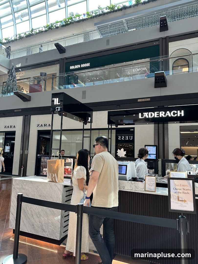 A view inside a multi-level shopping mall. In the foreground, a couple stands at a counter, possibly a food or retail outlet. Behind them, various shopfronts are visible, including Zara and Laderach. Further up, signs for Golden Goose and other brands can be seen. The mall features a modern design with a glass ceiling and decorative elements.