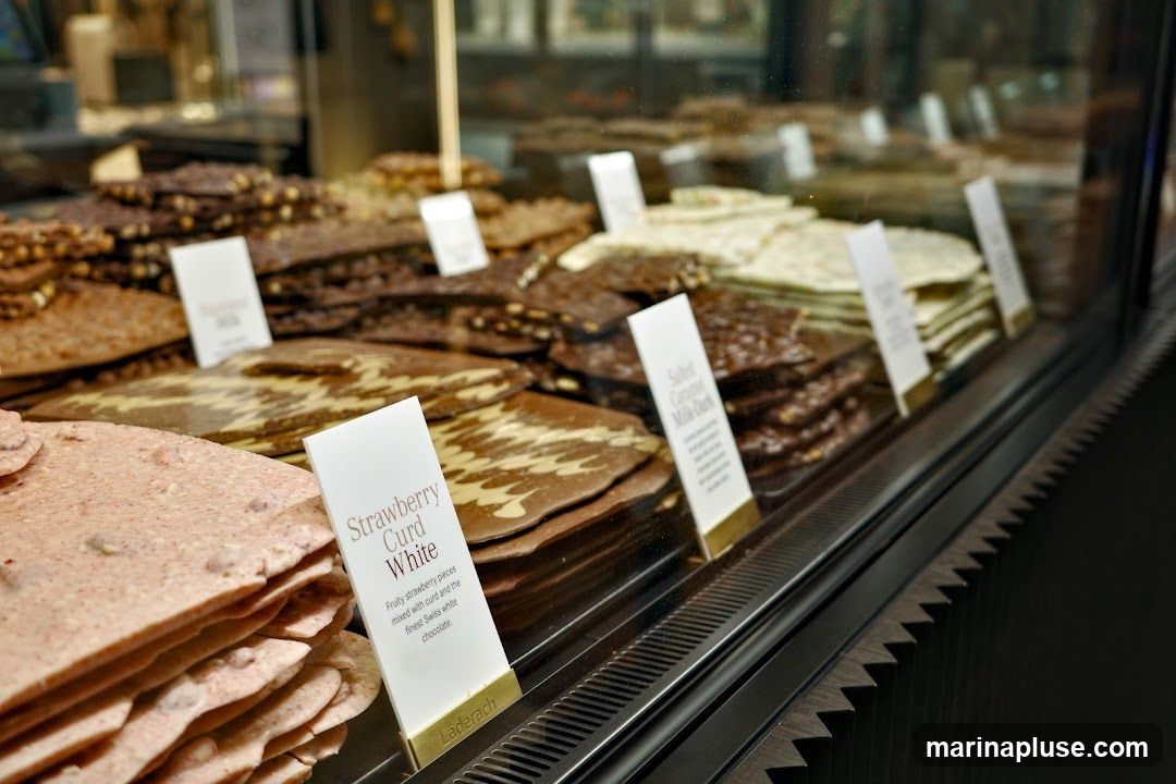 A display case filled with various types of chocolate bark. The chocolates are arranged in layers and have small white signs describing their flavors. Visible flavors include Strawberry Curd White and Salted Caramel Milk-Dark. The brand name "Läderach" is visible on one of the signs.