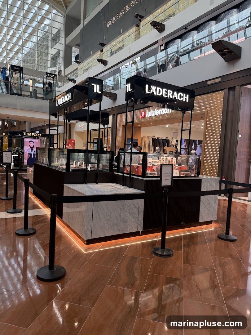 A Laderach chocolate kiosk is set up in a shopping mall. The kiosk has a modern design with black and marble accents. It is surrounded by stanchions and velvet ropes. In the background, other stores like Lululemon and Tom Ford are visible.