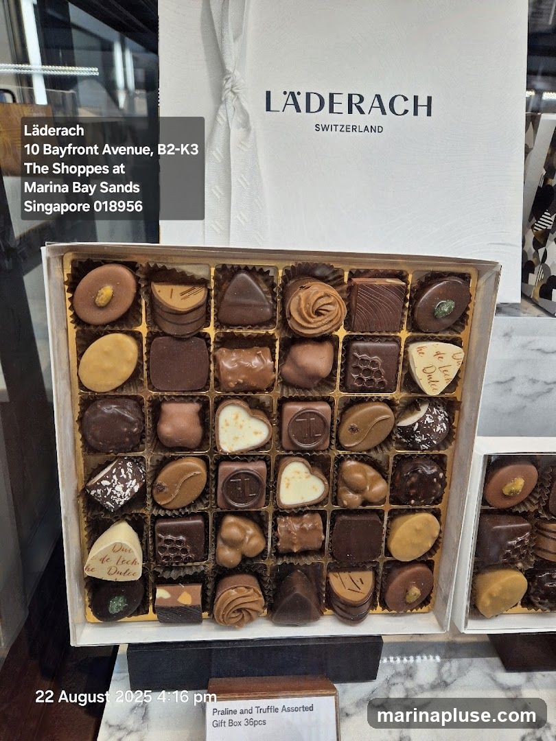 A close-up view of a gift box filled with assorted pralines and truffles from Läderach. The box contains a variety of chocolate shapes and designs, some with nuts and others with intricate patterns. The Läderach logo and store address are visible in the background. A label indicates it is a "Praline and Truffle Assorted Gift Box 36pcs".