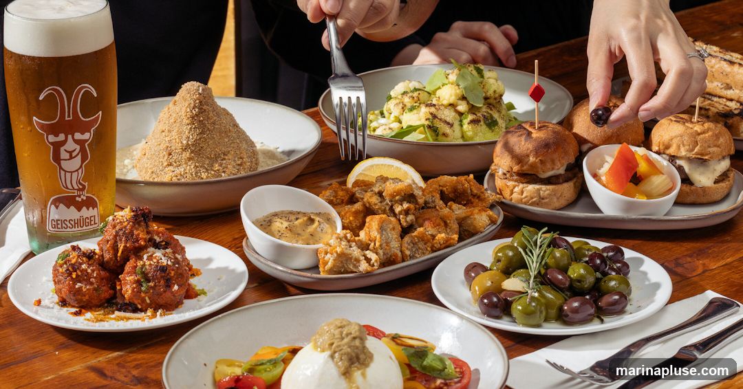 A table laden with various appetizers, including meatballs, fried bites, sliders, olives, and a salad, accompanied by a glass of beer with a goat logo and a dessert. Hands are visible reaching for the food.