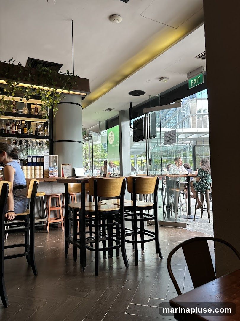 The image shows the interior of a bar or restaurant with a bar area on the left and seating areas visible through large glass windows. The bar is stocked with bottles and glassware. High chairs and tables are arranged throughout the space. People are seated at tables and at the bar. An "EXIT" sign is visible above the glass doors. A green sign with white text is visible through the glass.
