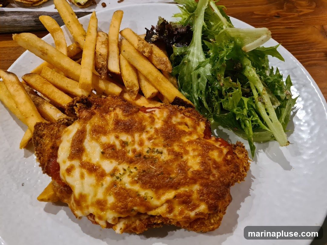 A close-up shot of a plate of food. The main dish appears to be chicken parmesan, a breaded chicken cutlet topped with marinara sauce and melted cheese, served with a side of golden french fries and a fresh green salad. The food is presented on a white, textured plate.