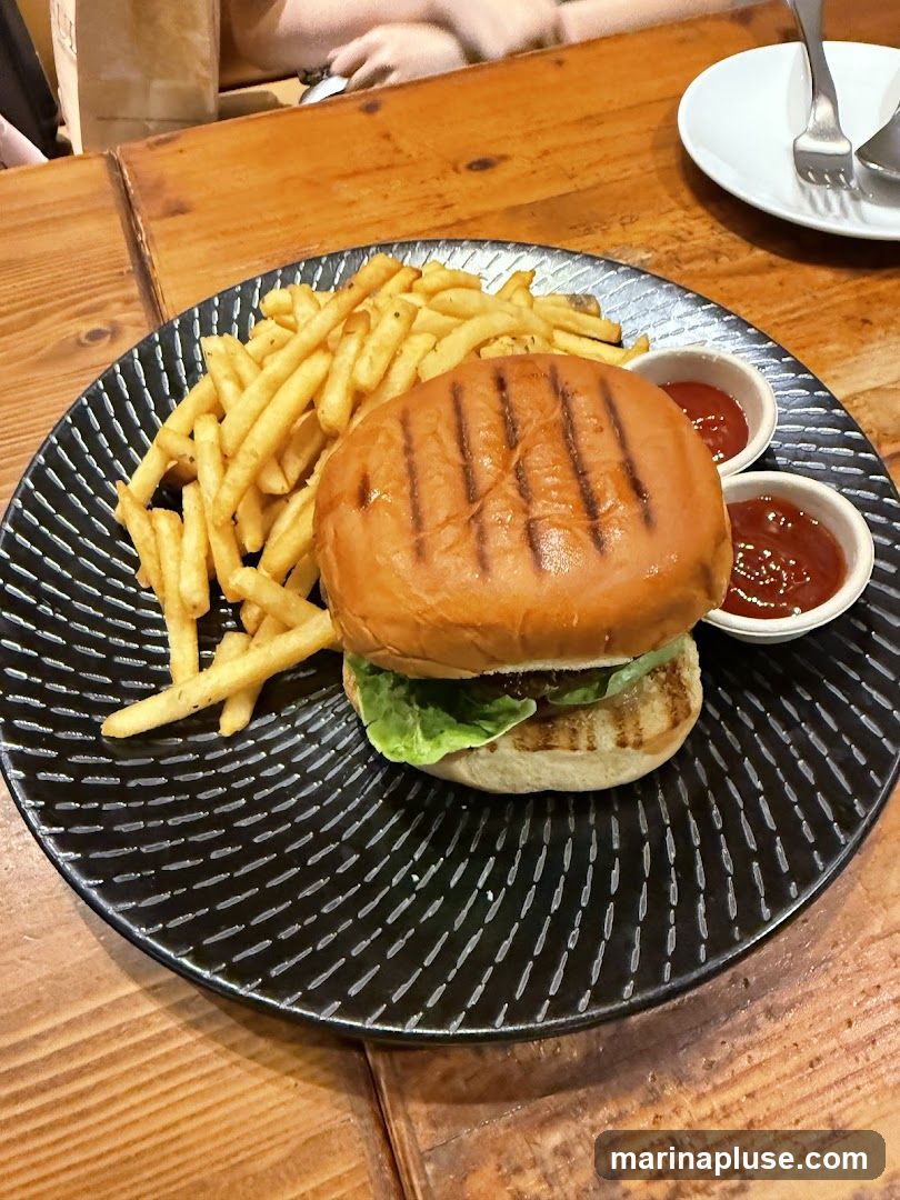 A close-up shot of a grilled burger with lettuce and two small bowls of ketchup, served with a side of french fries on a black patterned plate. The meal is placed on a wooden table.
