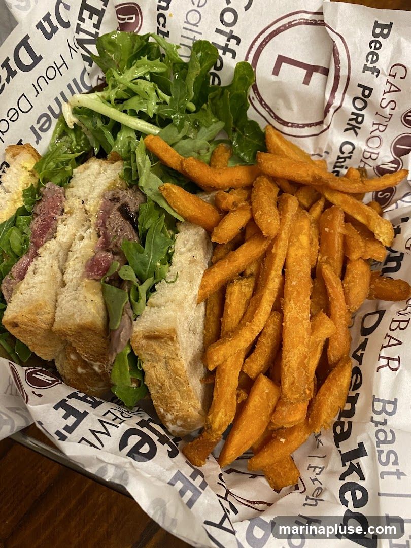A sandwich with sliced meat and greens, served with a side of sweet potato fries. The food is presented in a basket lined with paper featuring text and a logo.