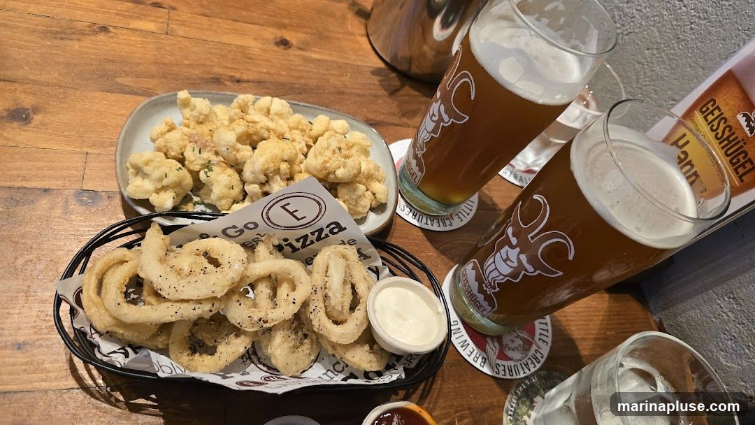 A table setting with two glasses of beer, a basket of fried calamari rings with a side of dipping sauce, and a plate of fried cauliflower. The beer glasses have logos for "Little Creatures Brewing" and "Geisshügel". A paper liner in the calamari basket has "Go E Pizza" printed on it.