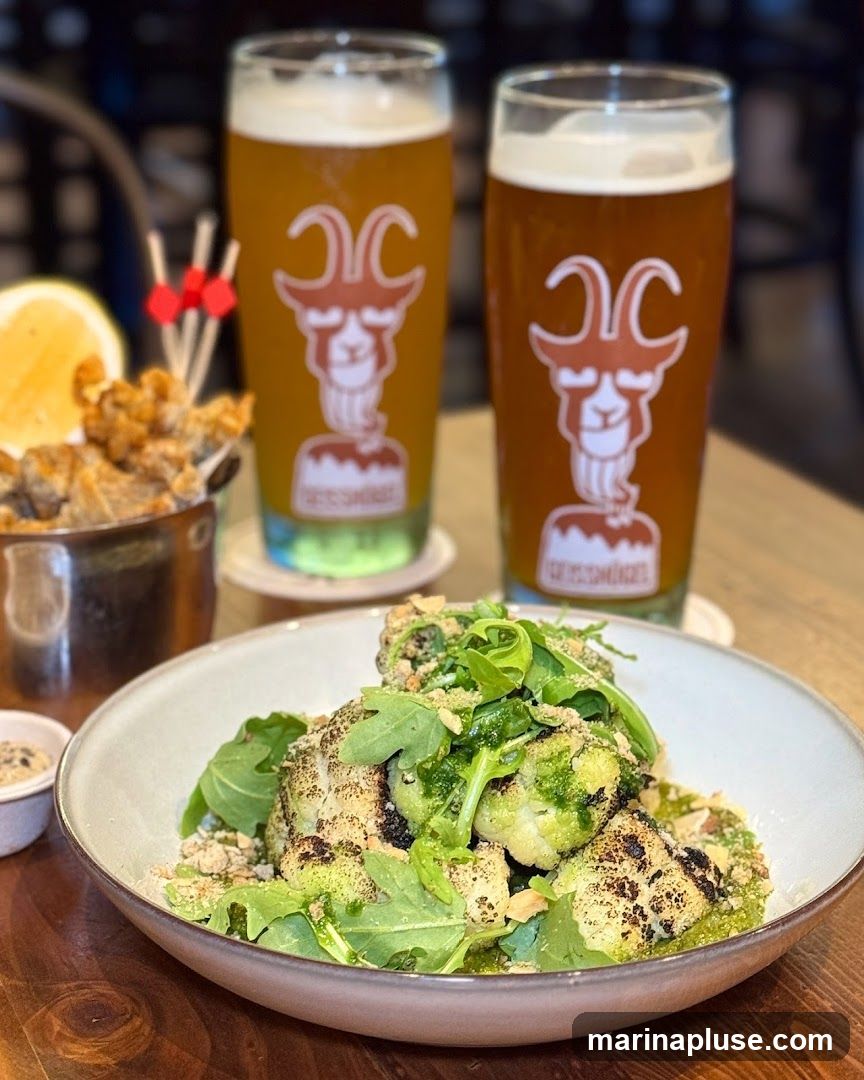 A close-up shot of a dish of roasted cauliflower topped with greens and nuts, served in a bowl. To the left, a metal container holds fried appetizers, with a lemon slice and toothpicks with red bows. In the background, two glasses of beer are visible, each featuring a logo of a goat's head.