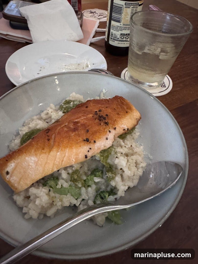 A close-up shot of a dish featuring a piece of pan-seared salmon atop a creamy risotto with broccoli. A fork and spoon are visible, along with a glass of a light-colored beverage, a coaster, a used plate, and a bottle in the background.