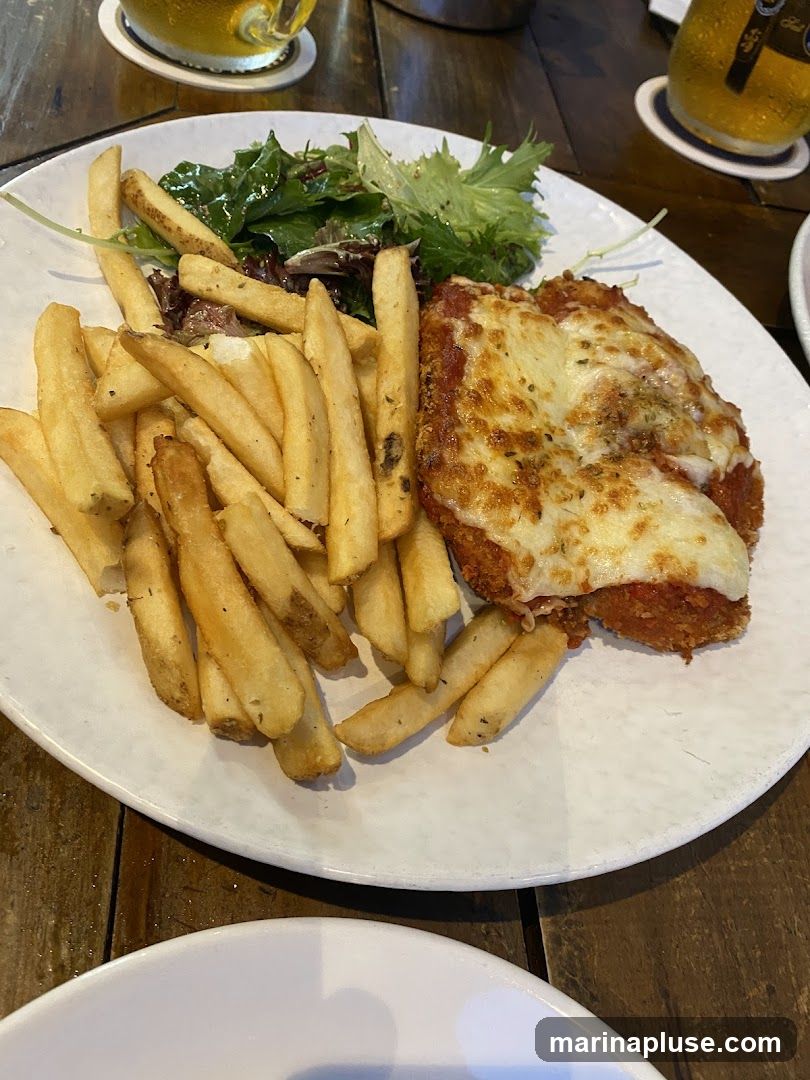 A plate of chicken parmesan topped with melted cheese and tomato sauce, served with a side of french fries and a small green salad. Two glasses of beer are visible in the background.