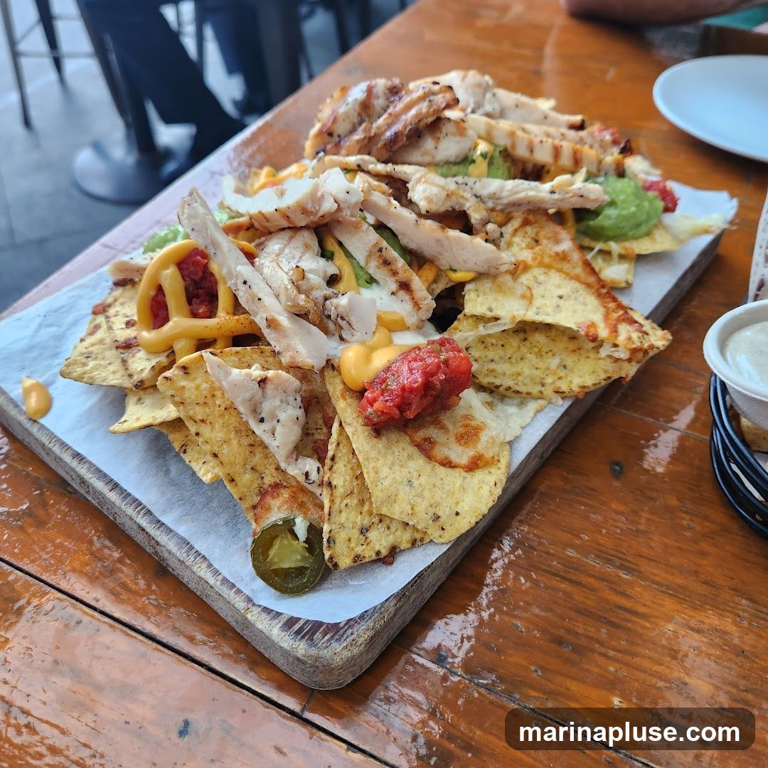 A platter of nachos topped with grilled chicken, guacamole, cheese sauce, and salsa, served on a wooden board with parchment paper. A jalapeño slice is visible on the side.