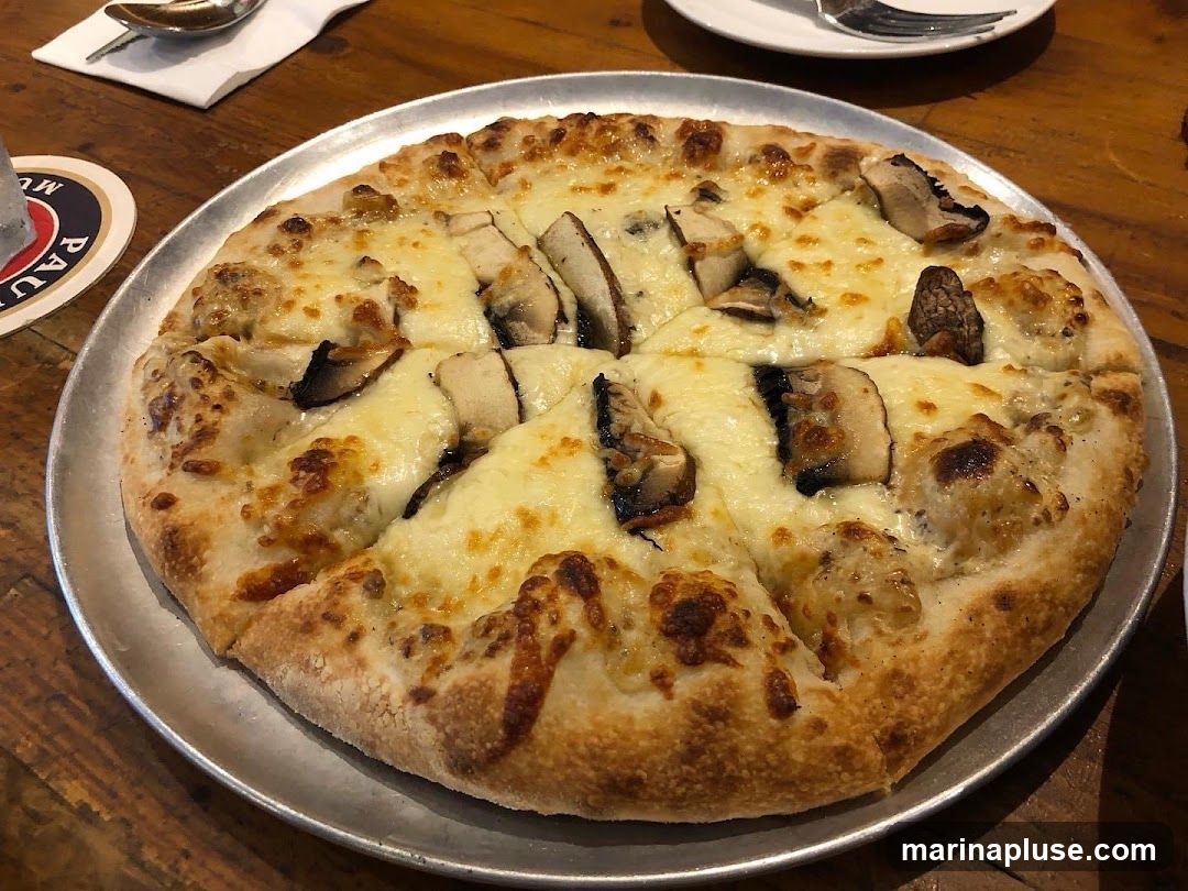 A close-up shot of a mushroom pizza on a metal platter, placed on a wooden table. The pizza has a golden-brown crust, melted cheese, and slices of mushrooms. A coaster with the word "Paul" is visible on the left side of the image.