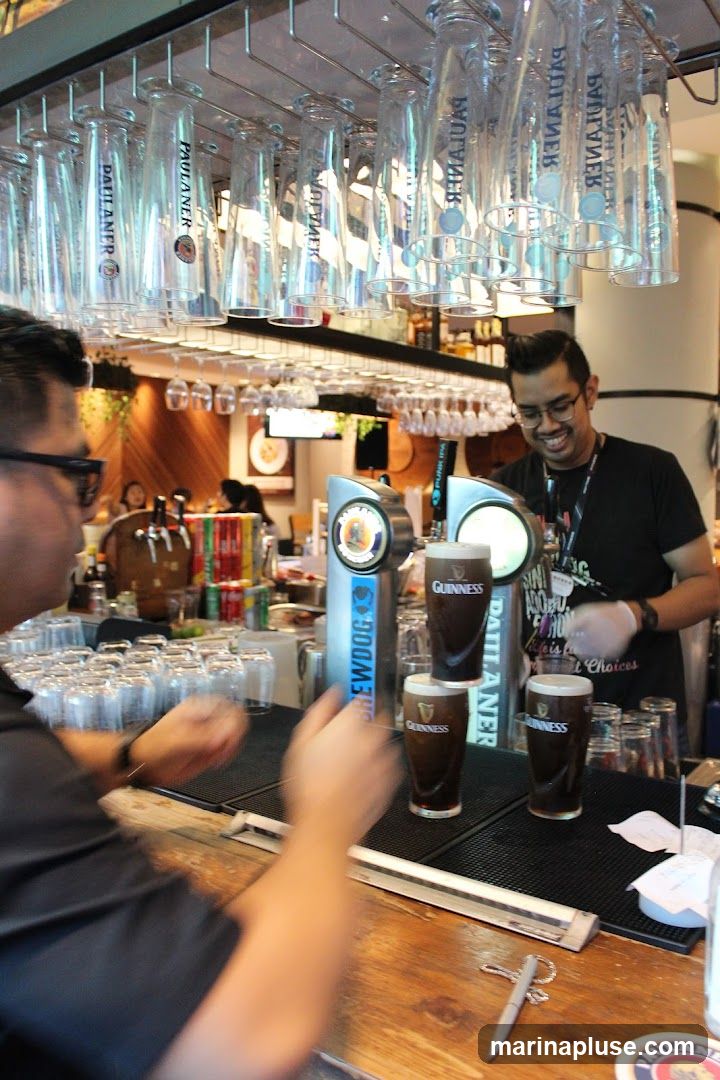 A bartender wearing gloves and a black t-shirt smiles as he pours a Guinness beer at a bar. Several glasses of Guinness are stacked and lined up on the bar. Many empty glasses are hanging upside down above the bar. In the background, there are shelves stocked with bottles and cans.