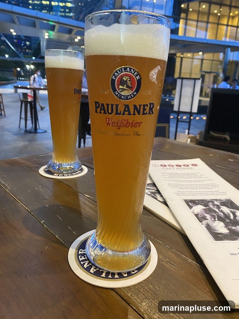 Two tall glasses of Paulaner Weissbier, a German wheat beer, are placed on a wooden table. The glasses are filled with golden beer and topped with a frothy white head. The glass in the foreground is larger and shows the Paulaner logo prominently. Coasters with the Paulaner logo are under each glass. In the background, a blurred view of a modern city skyline and some patrons can be seen. A menu is also visible on the table.